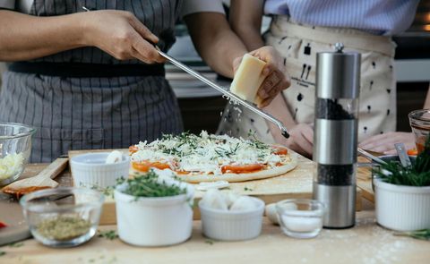 Two people grate cheese onto a pizza, kitchen setting.