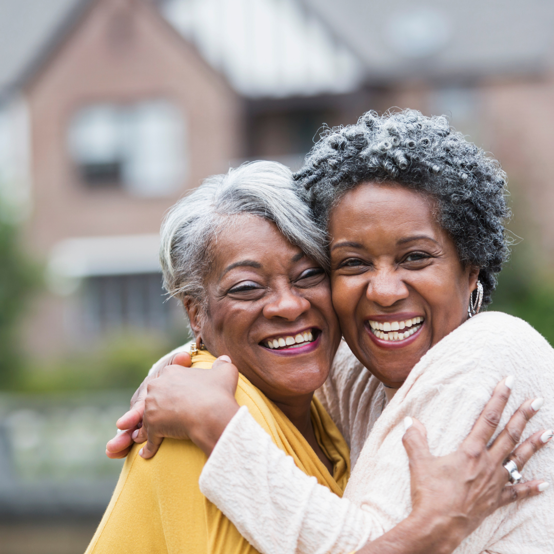 Two older women are hugging each other and smiling in front of a house.