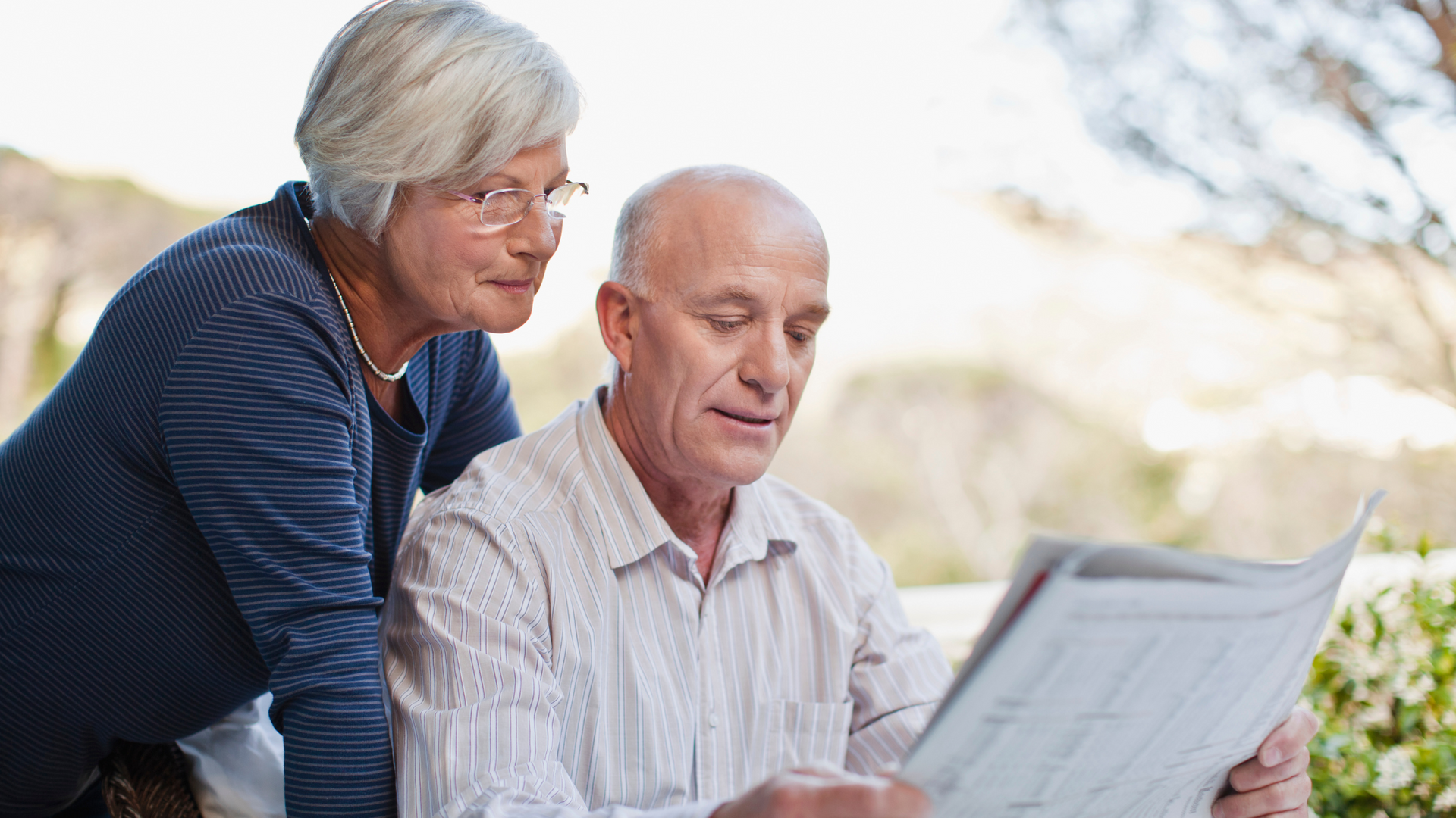 Older couple reading newspaper together outdoors; woman leans over man's shoulder, both focused.