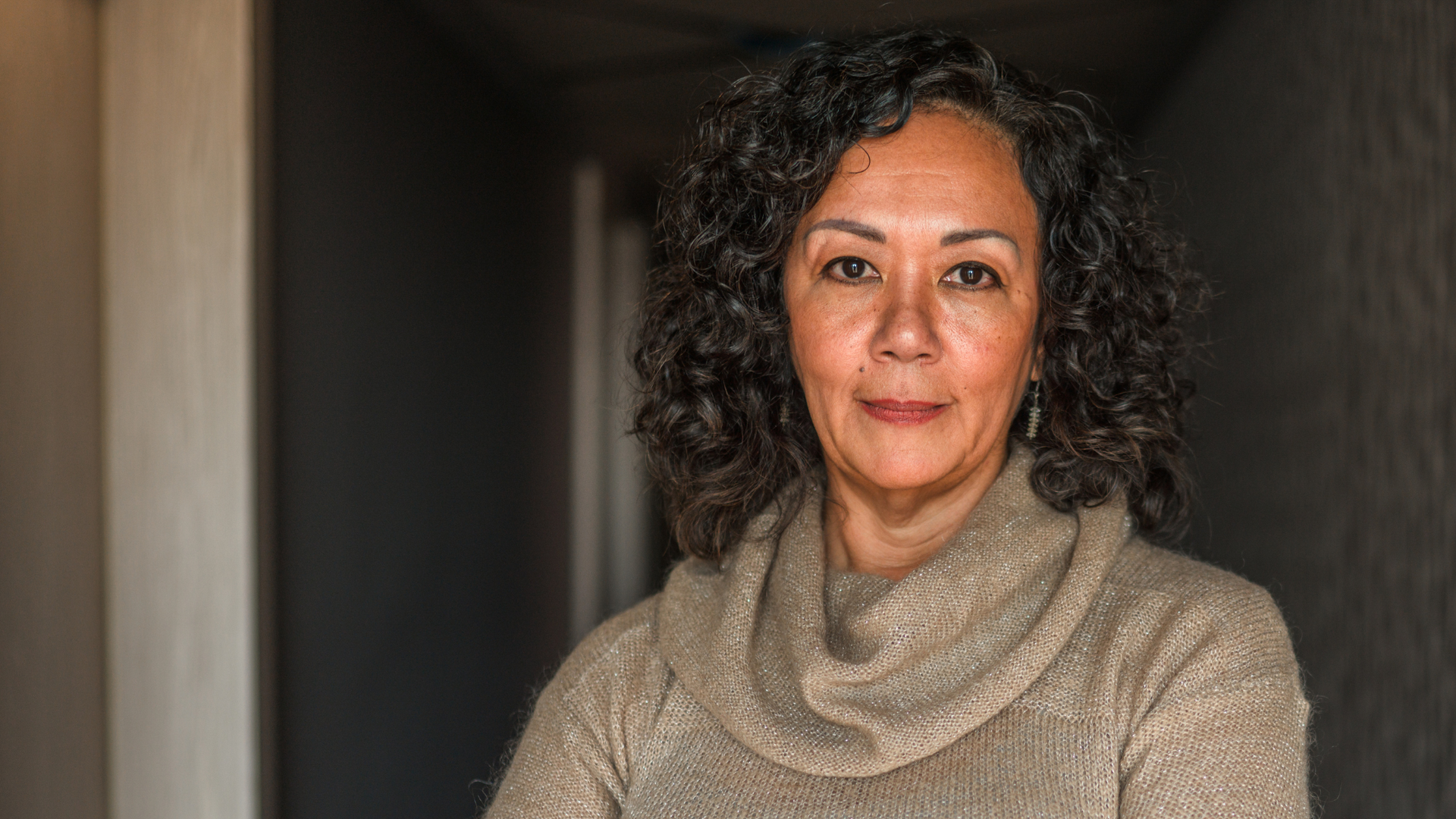 A professional woman with curly hair is wearing a sweater and standing in a hallway.
