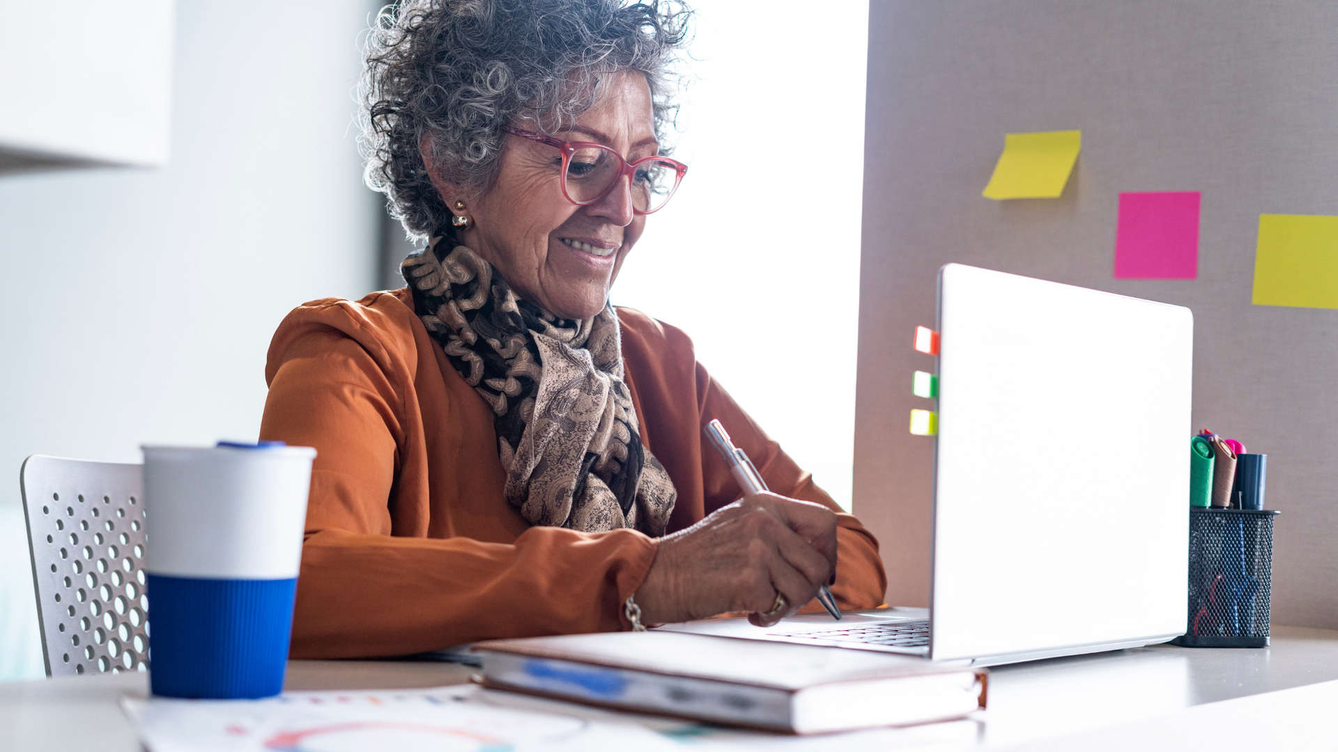 An elderly woman is sitting at a desk using a laptop computer.