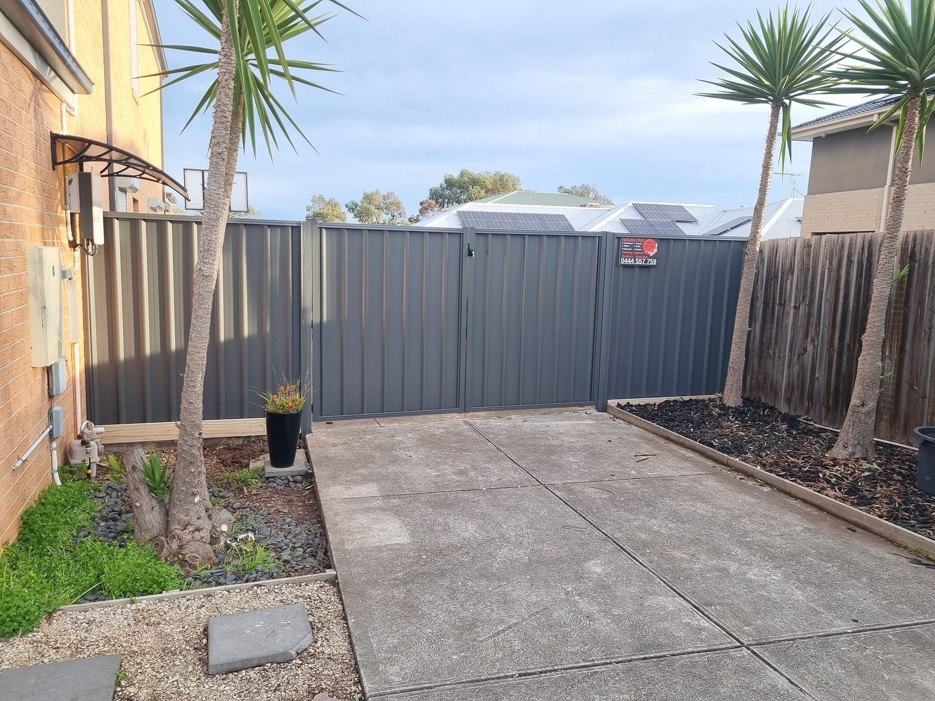 Gray metal fence with gate, concrete driveway, palm trees, and sky.