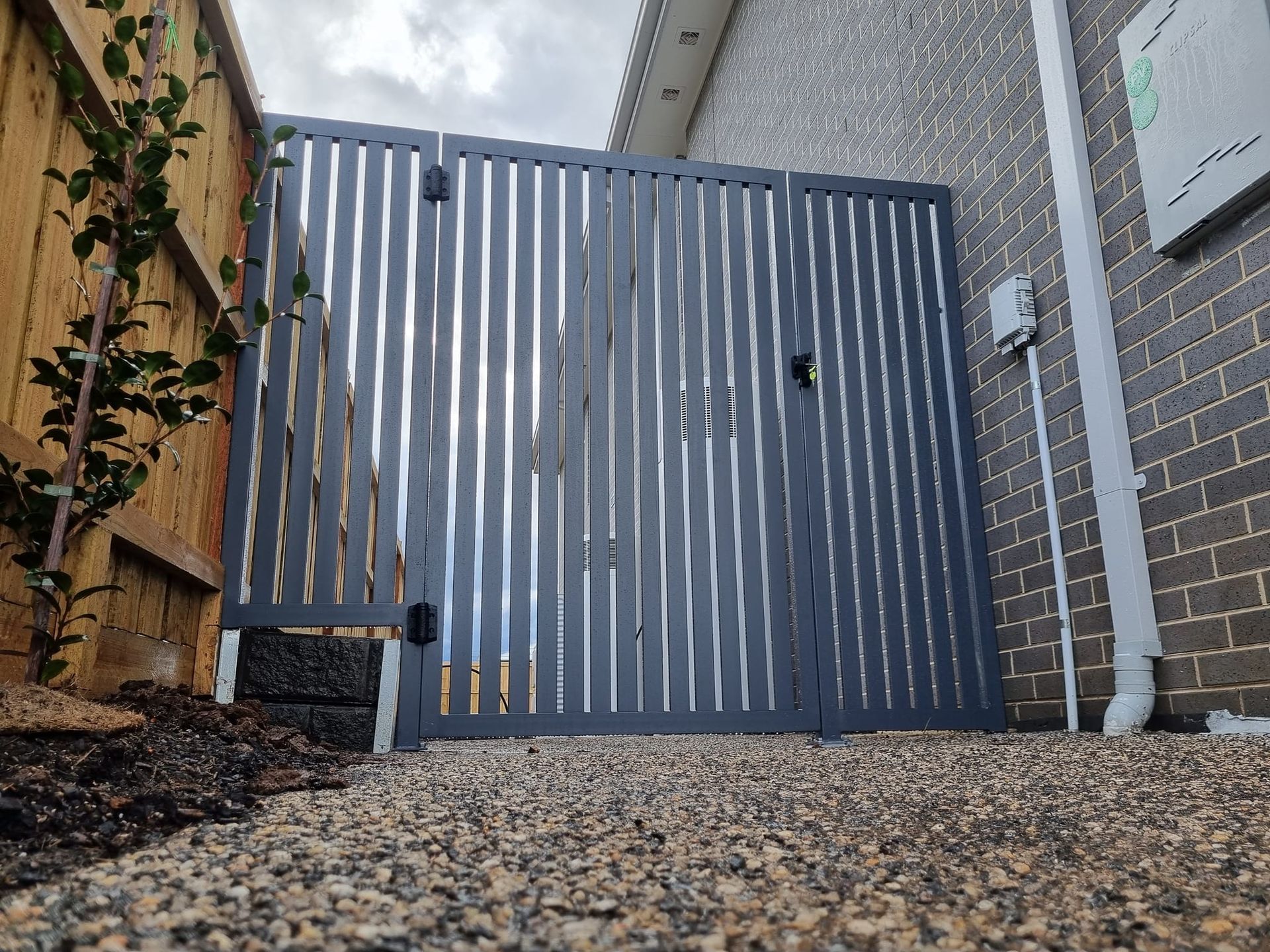Dark gray slat gate between a wooden fence and a brick wall, gravel ground.
