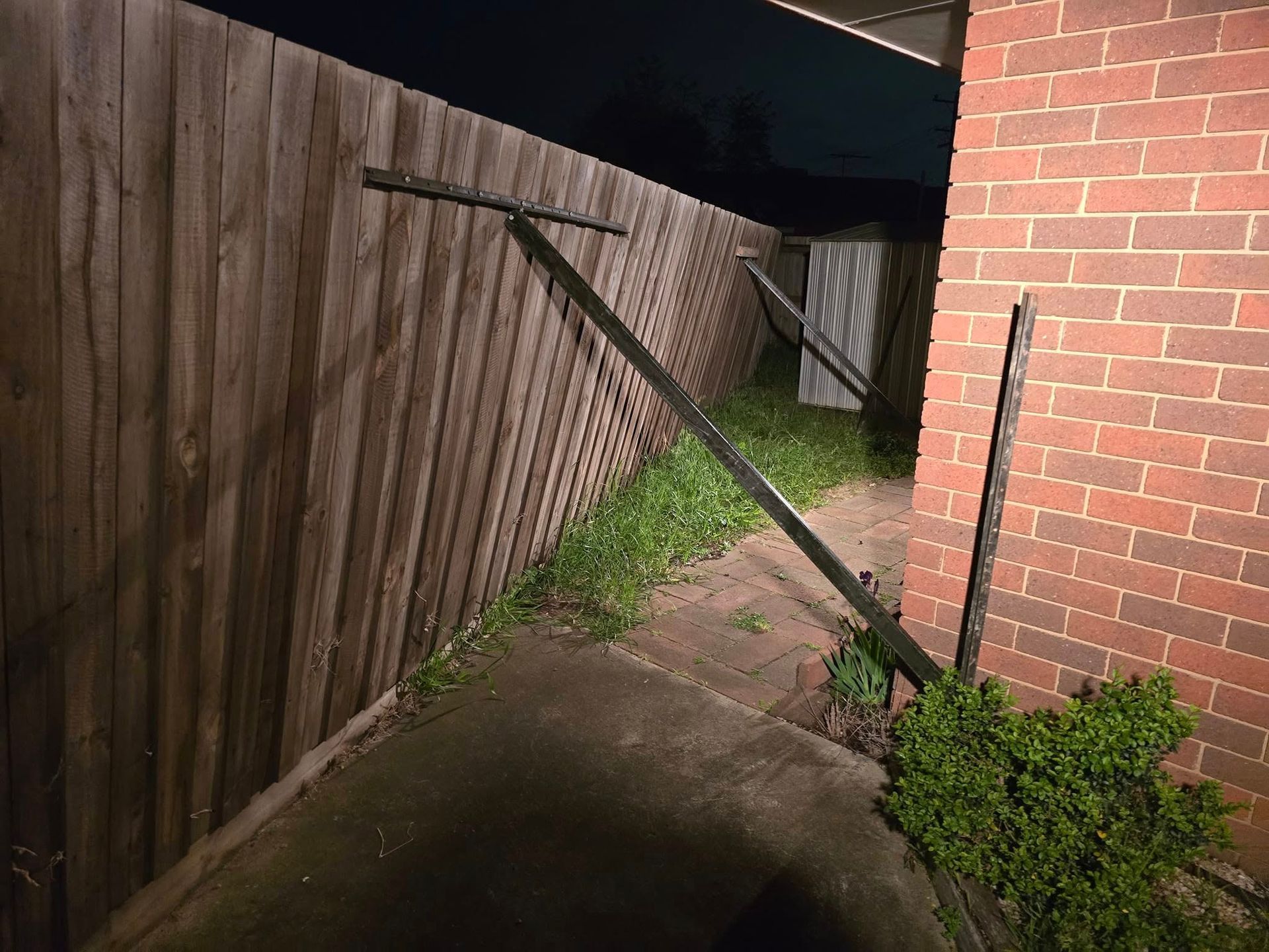Wooden fence leaning at an angle, damaged with metal beam against it, near a brick wall and sidewalk.