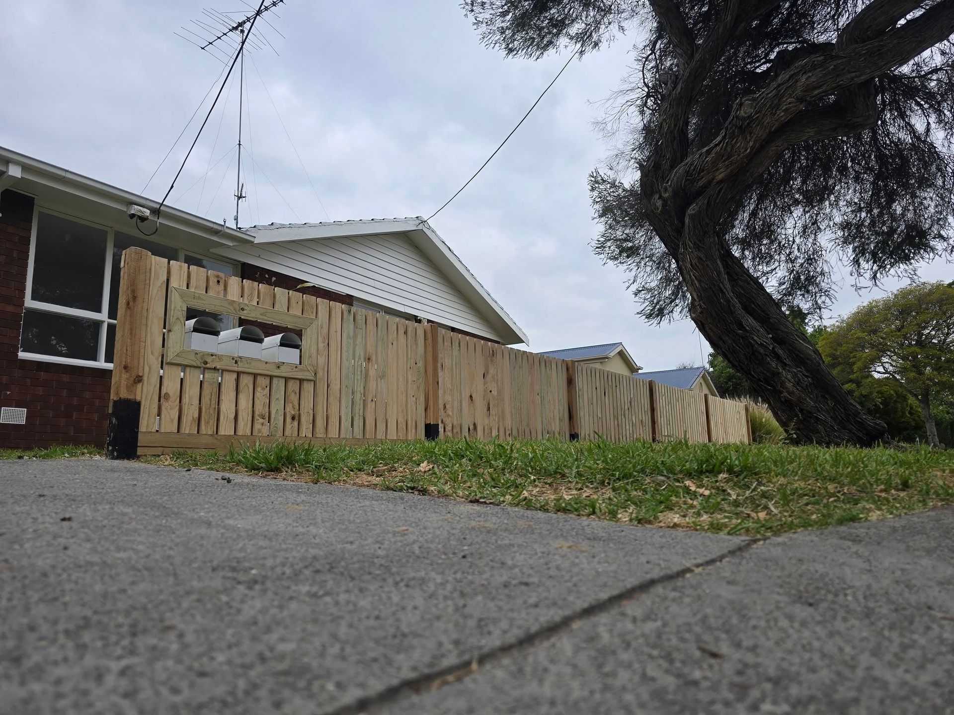 Wooden fence in front of a house, a tree to the right, and a cloudy sky.