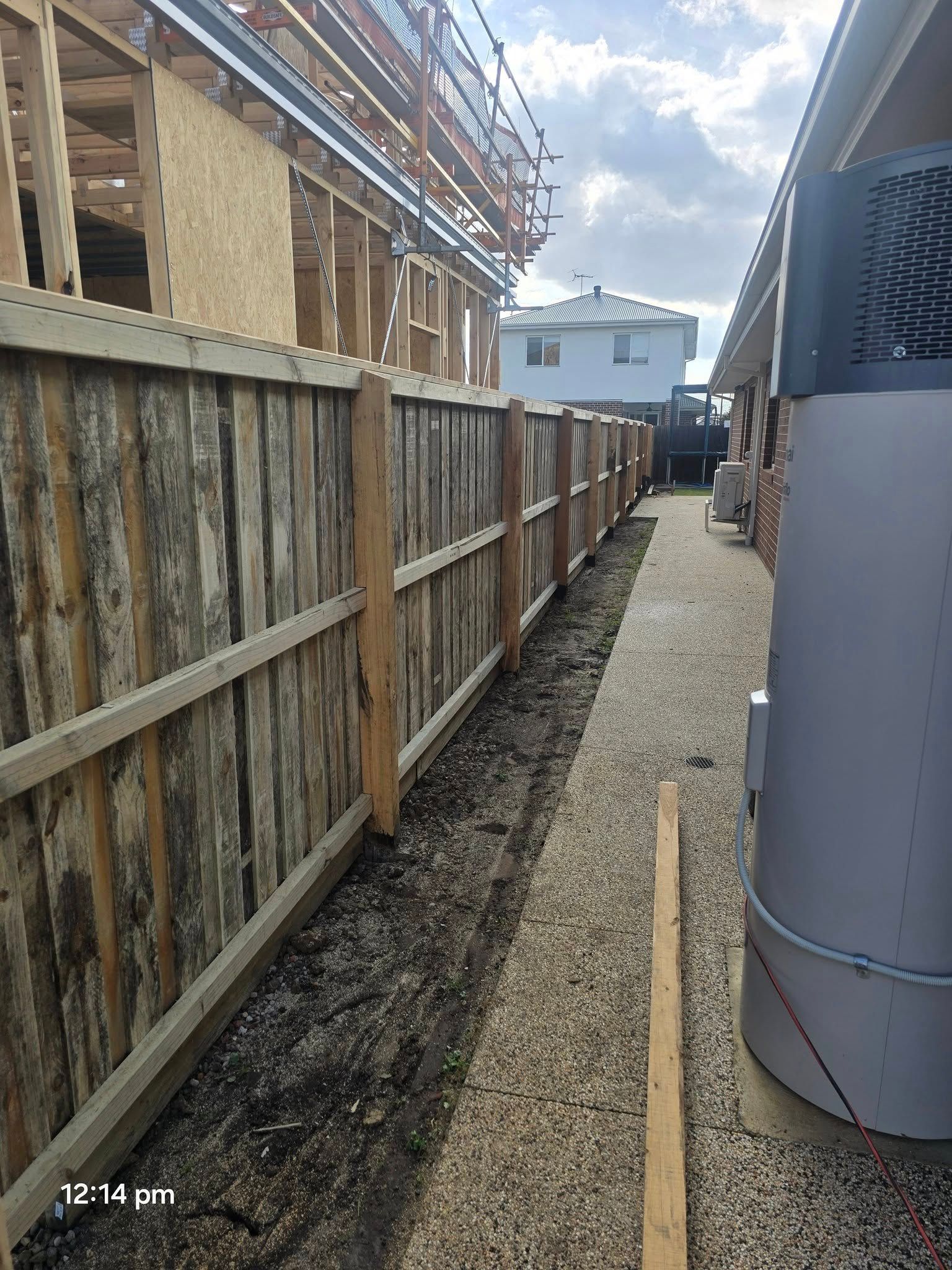 A wooden fence runs along a gravel path between two buildings under a cloudy sky.