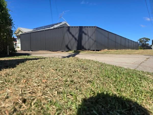 A long, dark gray corrugated metal fence along a street, with a grassy lawn in the foreground and a house visible behind it.