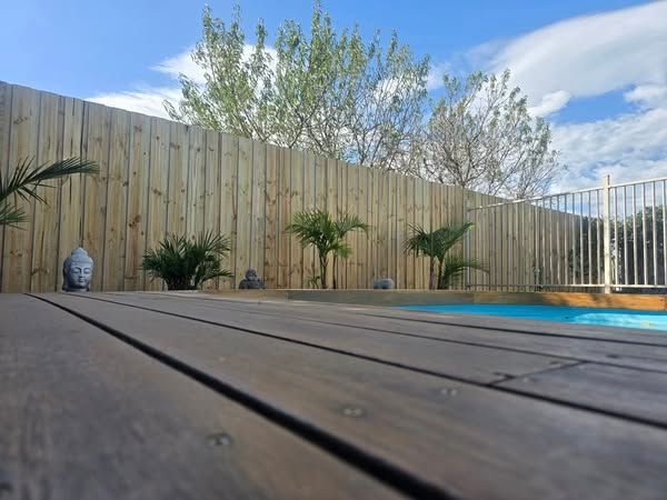 Wooden deck and fence surround a pool with palm trees, a Buddha statue, and a blue sky.