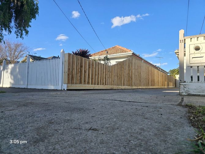A concrete driveway leads to a long wooden fence. Bright blue sky with few clouds.