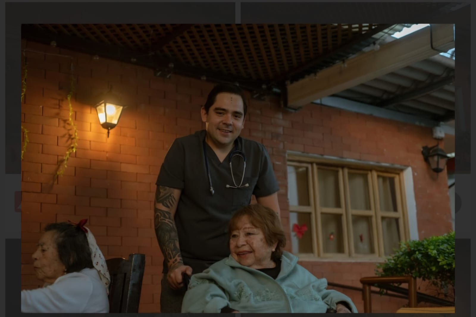 Un hombre de pie detrás de una mujer sentada en un patio de ladrillos al atardecer, con la cálida luz de una linterna