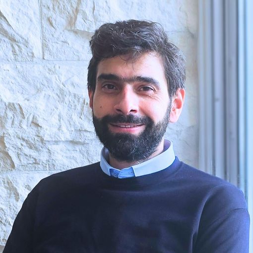 Man in a blazer gestures with hands, seated near a window and stone wall, smiling.