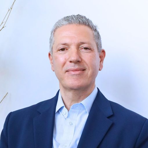 Man in tan jacket, looking at the camera, against a textured stone wall background.