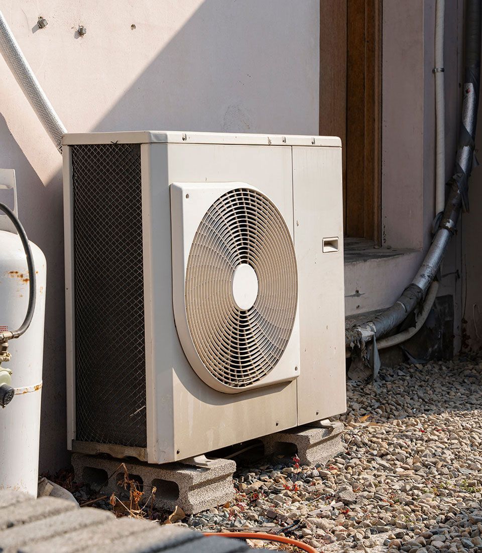 An outdoor air conditioning unit sits on cinder blocks. A pipe runs down the exterior wall near the door