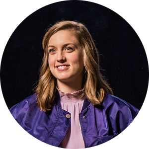 Woman with brown wavy hair smiles, wearing a black blazer and white shirt, in a warm-toned room.