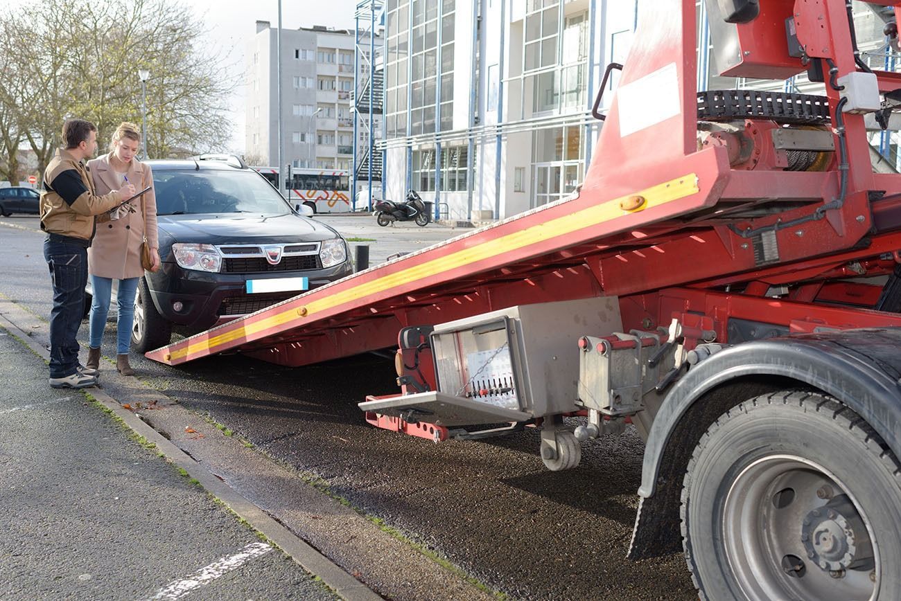 A man and woman are standing next to a tow truck with a car on the back.