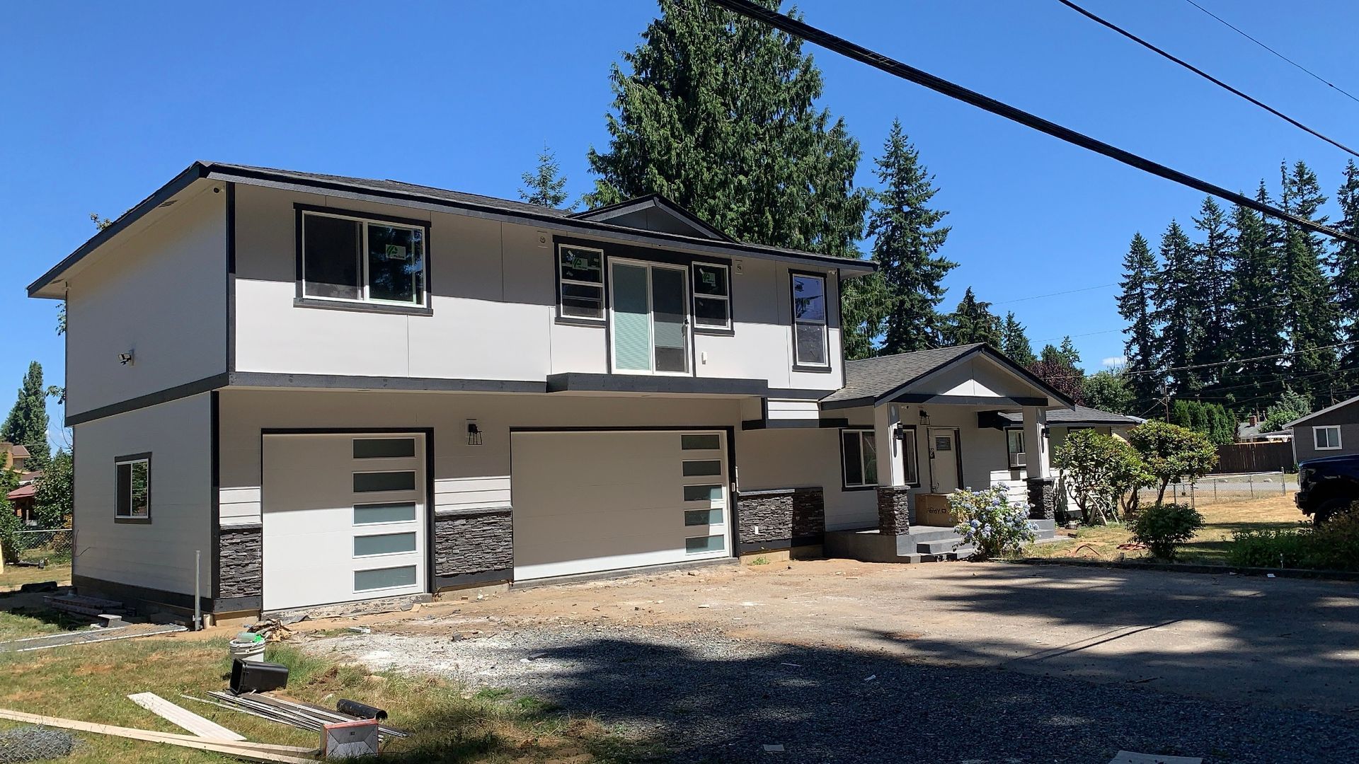 Two-story house with gray stucco, black roof, and modern garage doors. Gravel driveway under blue sky.