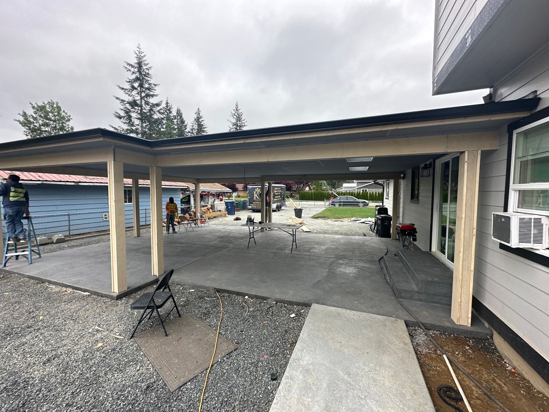 A covered outdoor patio under construction; workers on a ladder. Gray concrete, gravel ground, overcast sky.