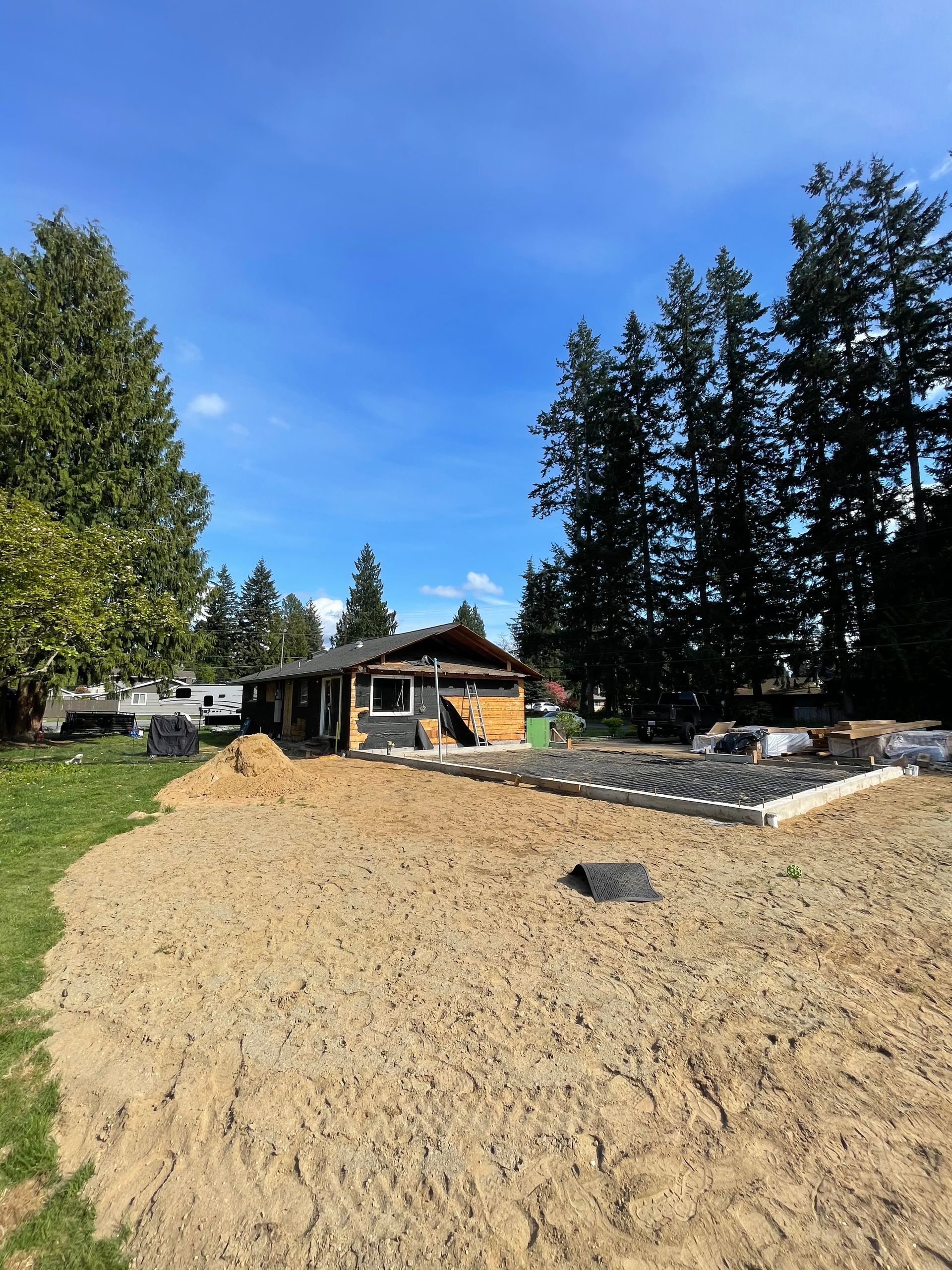 House under construction with exposed framing, set on a sandy lot, under a blue sky.