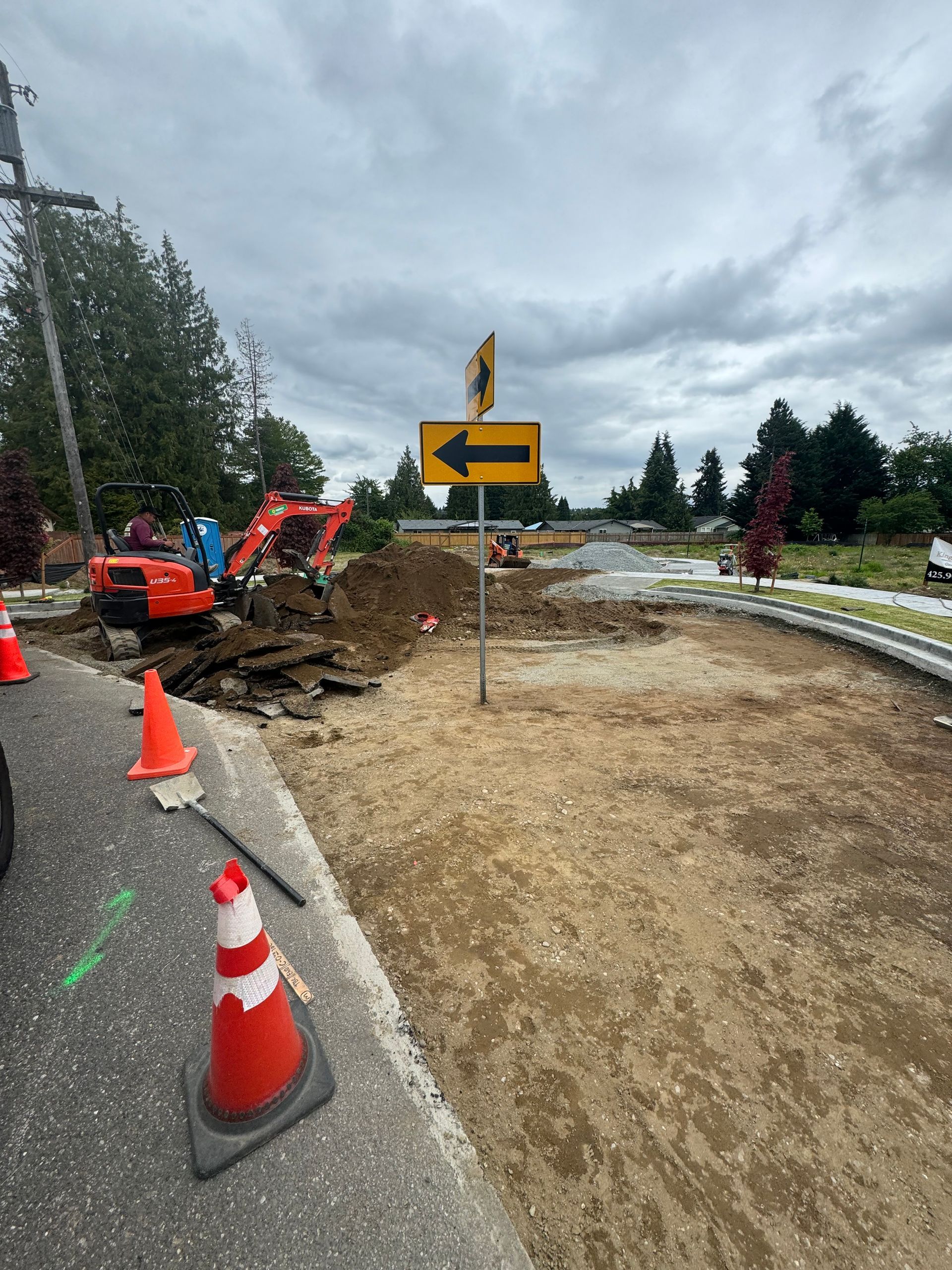 Road construction at a roundabout. Two excavators dig, traffic cones present. Cloudy sky.