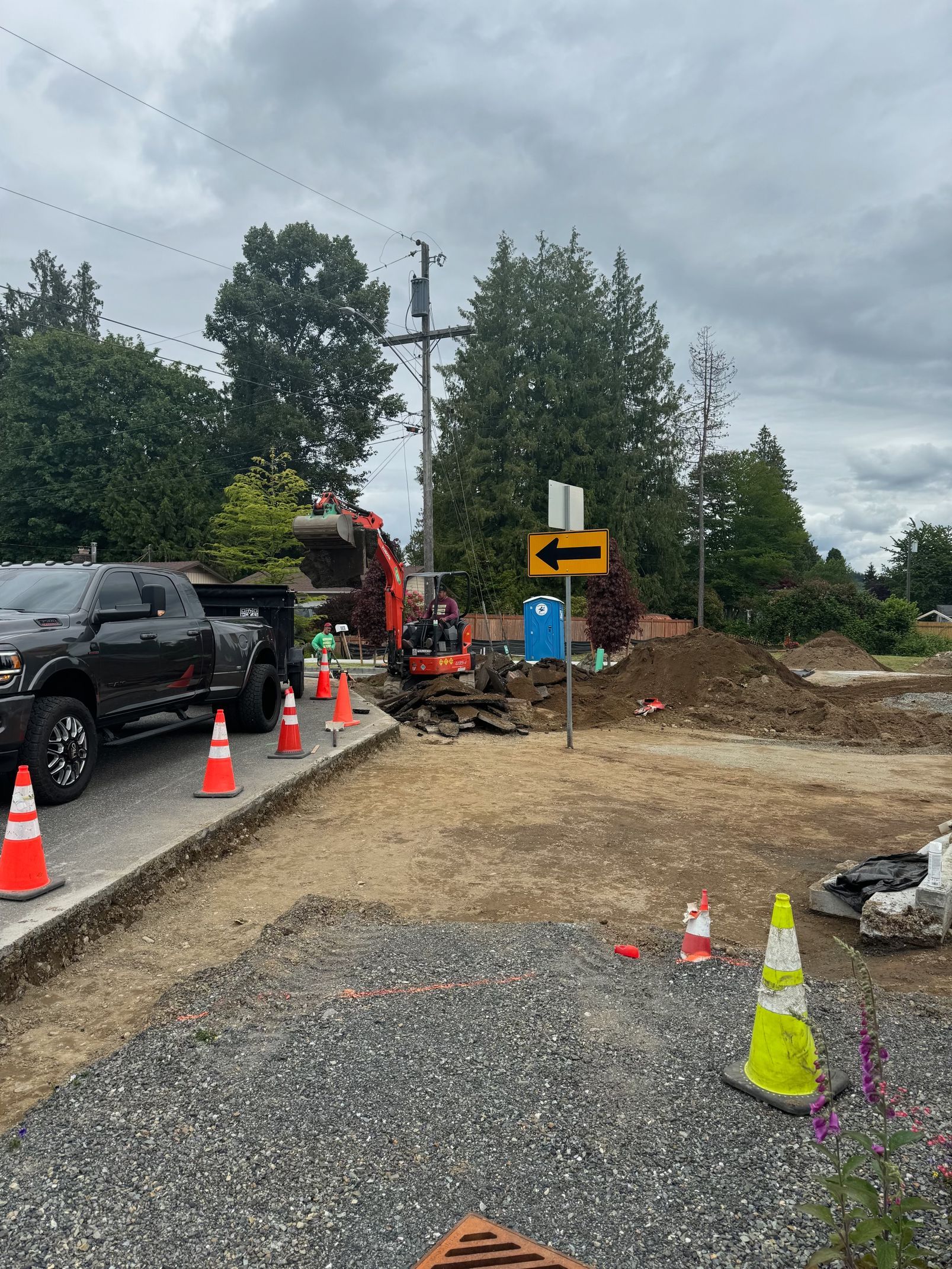 Construction site with excavator and truck. Orange cones, dirt pile, and sign. Cloudy day.