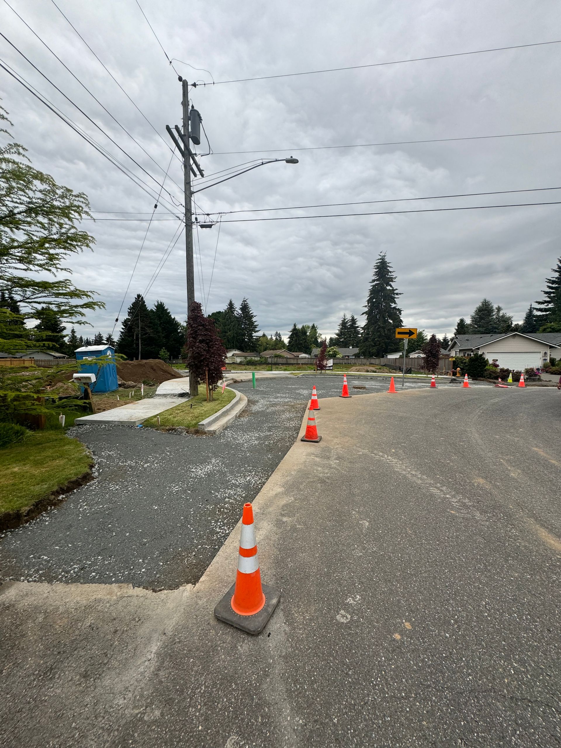 Road under construction with orange cones, gravel, and utility pole, under cloudy sky.