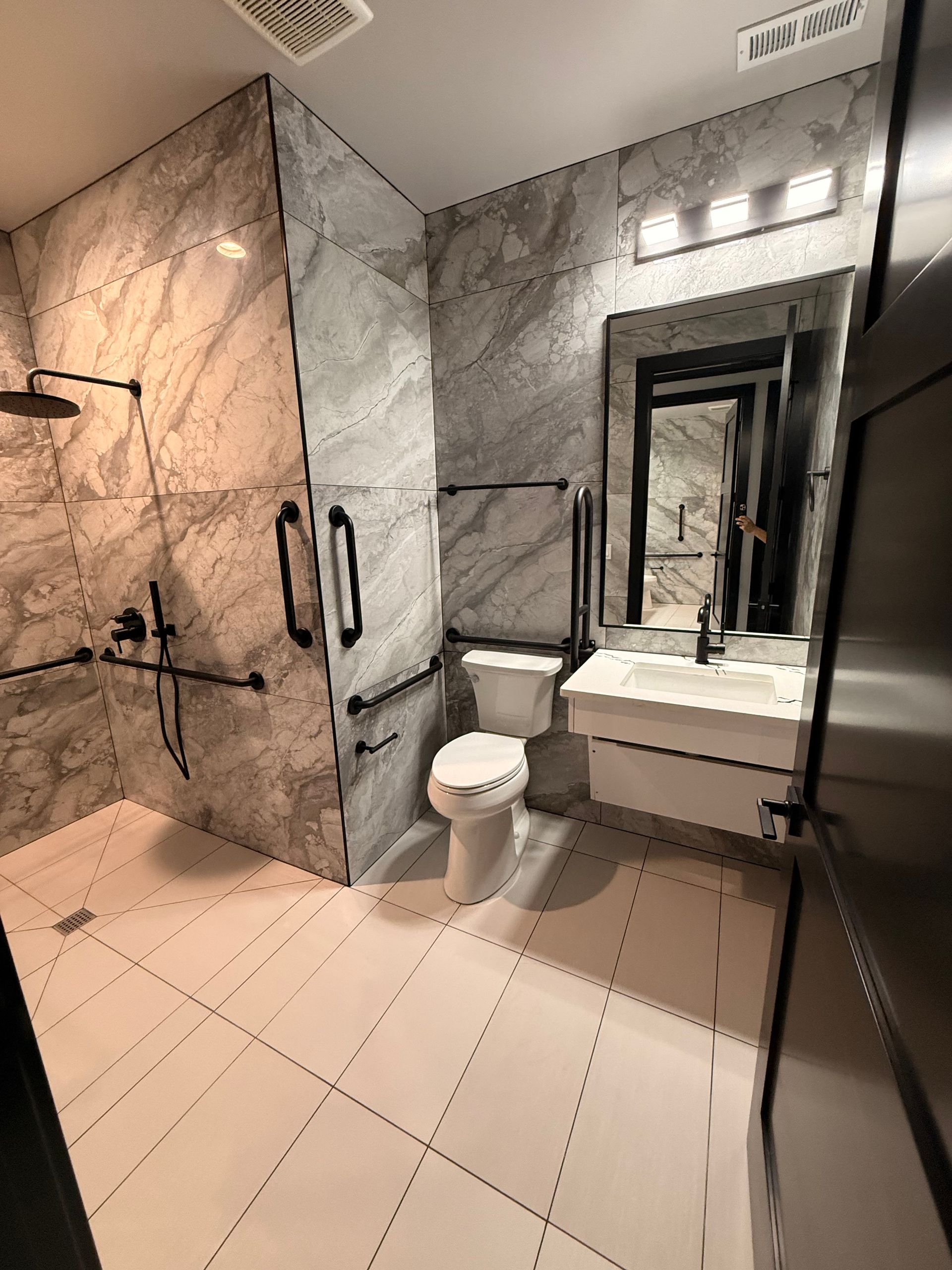 Modern bathroom with marble walls, accessible shower, toilet, sink, and mirror.