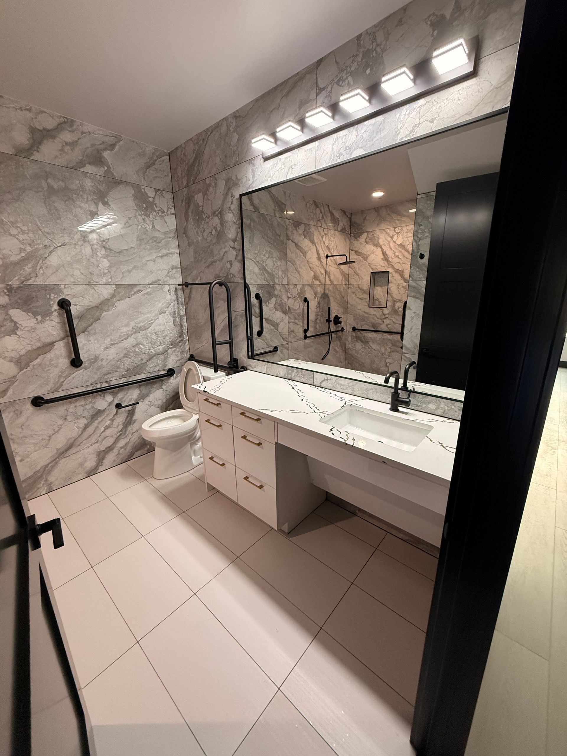 Modern accessible bathroom with marble walls, large mirror, and grab bars. White sink and toilet.