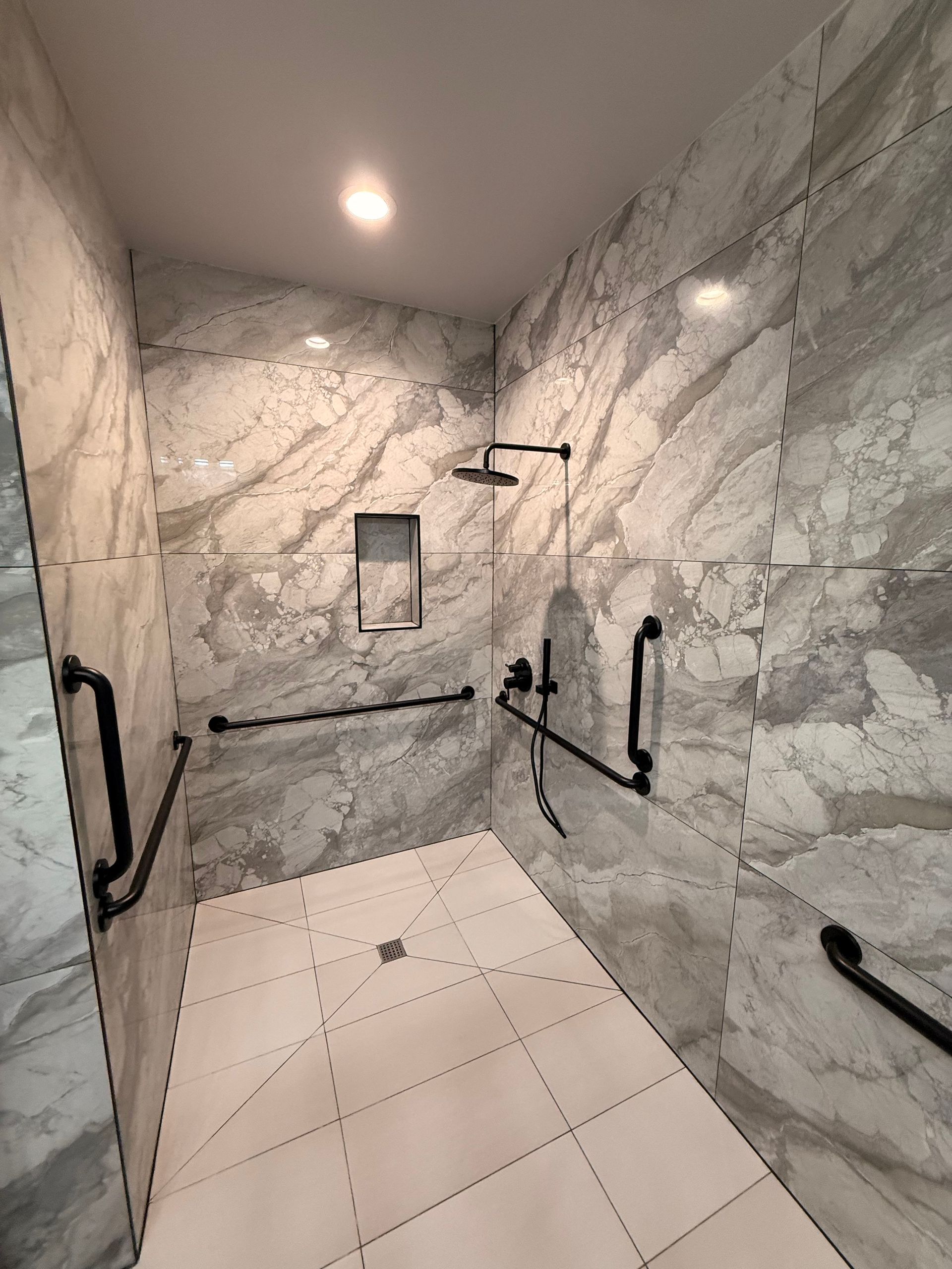 Accessible shower with marble-like gray tile, black grab bars, showerhead, and a recessed soap niche.