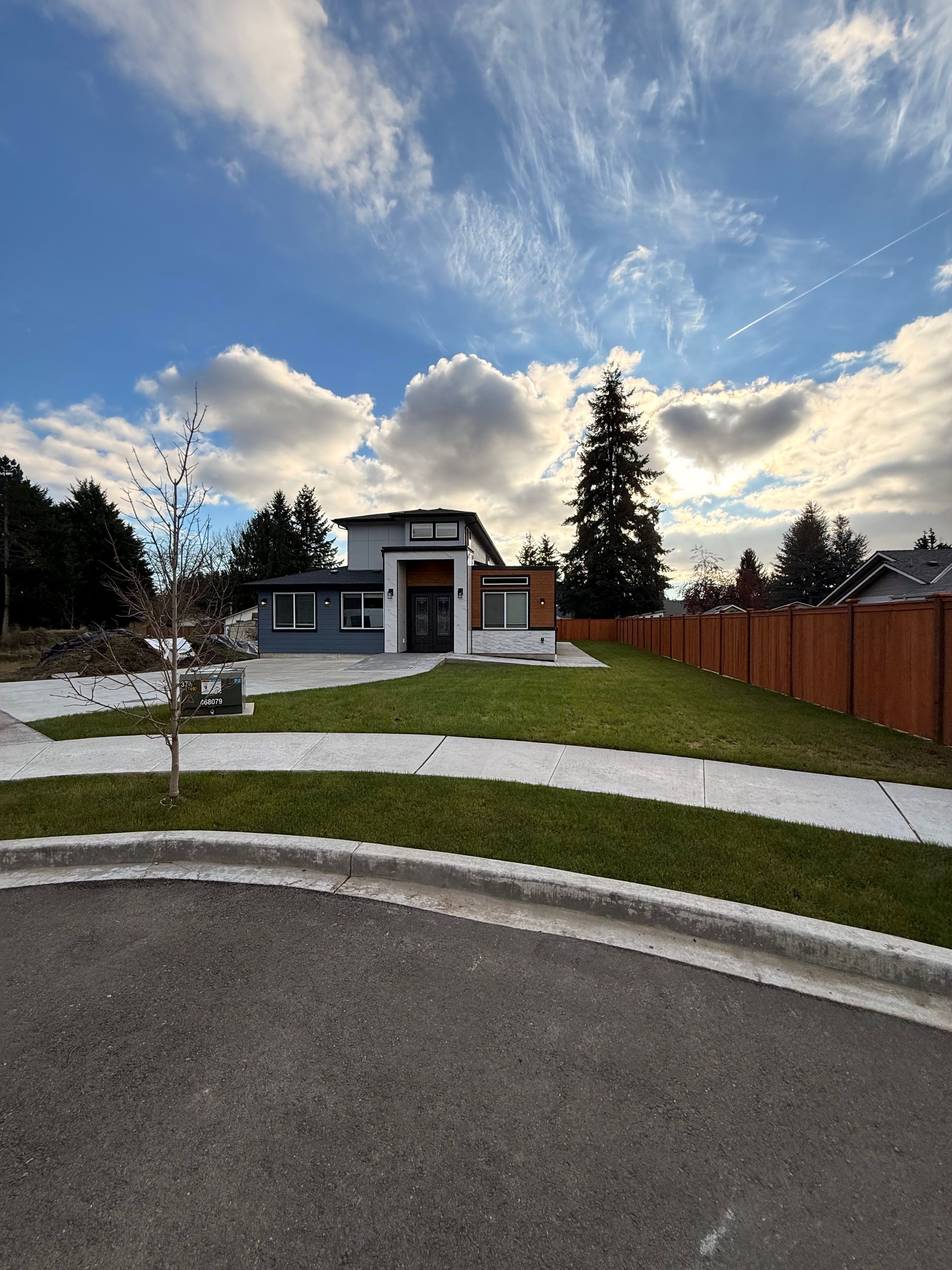 Modern house with gray and white facade, green lawn, blue sky, and brown fence.