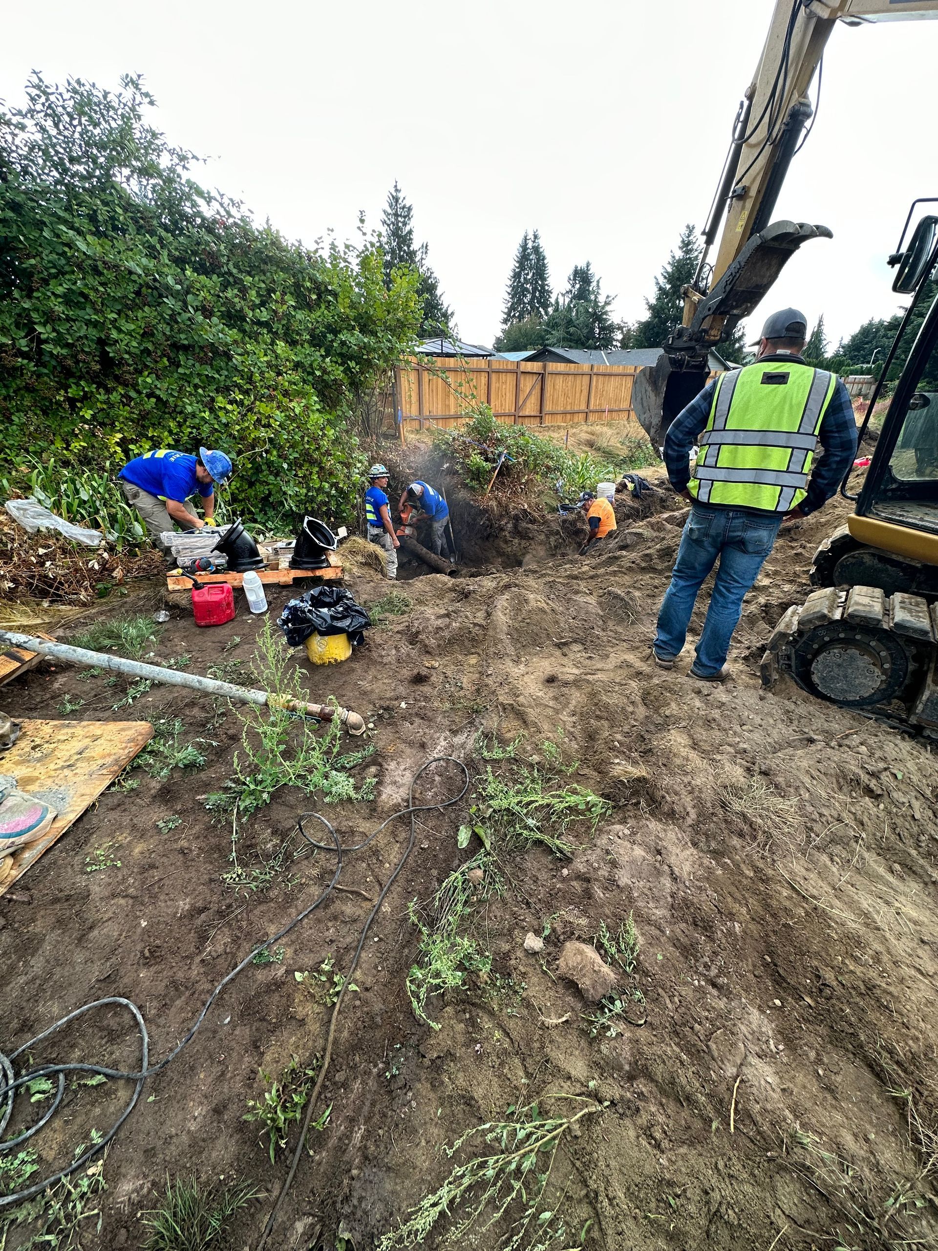Construction workers excavating a muddy area with an excavator, working near trees and a fence.