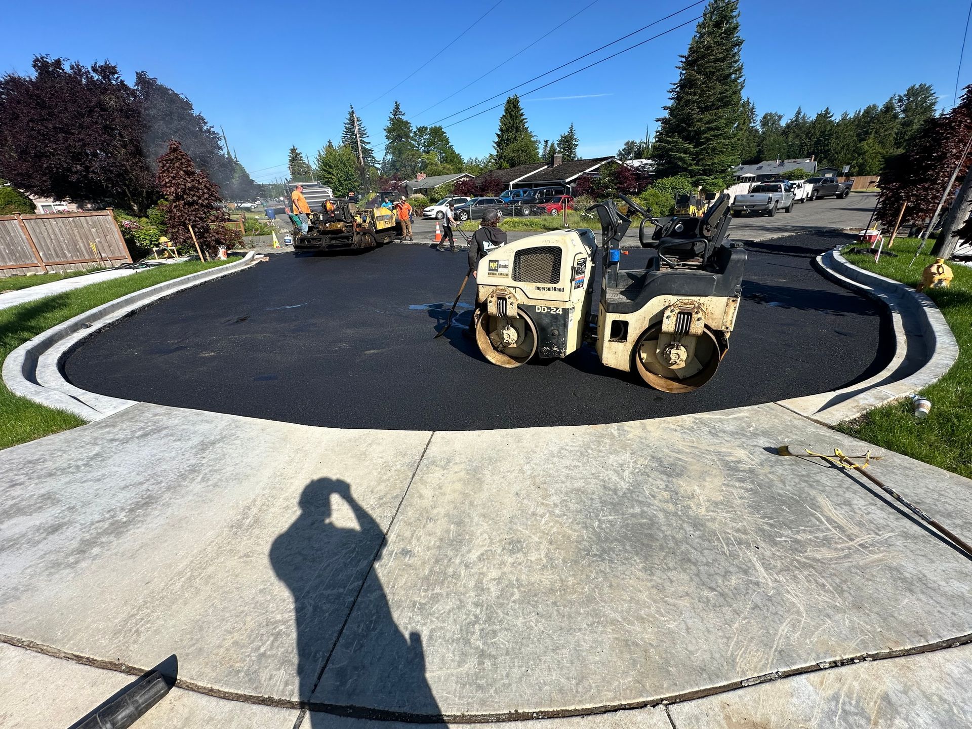 Asphalt paving of a residential driveway, with workers, rollers, and vehicles in the background.