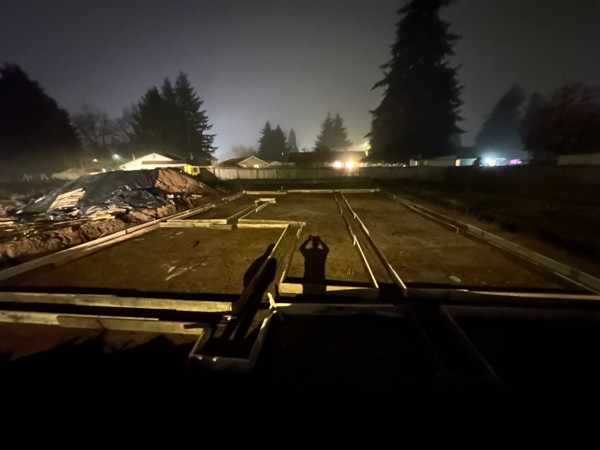 Construction site at night with a level foundation, wooden forms, and two people visible.