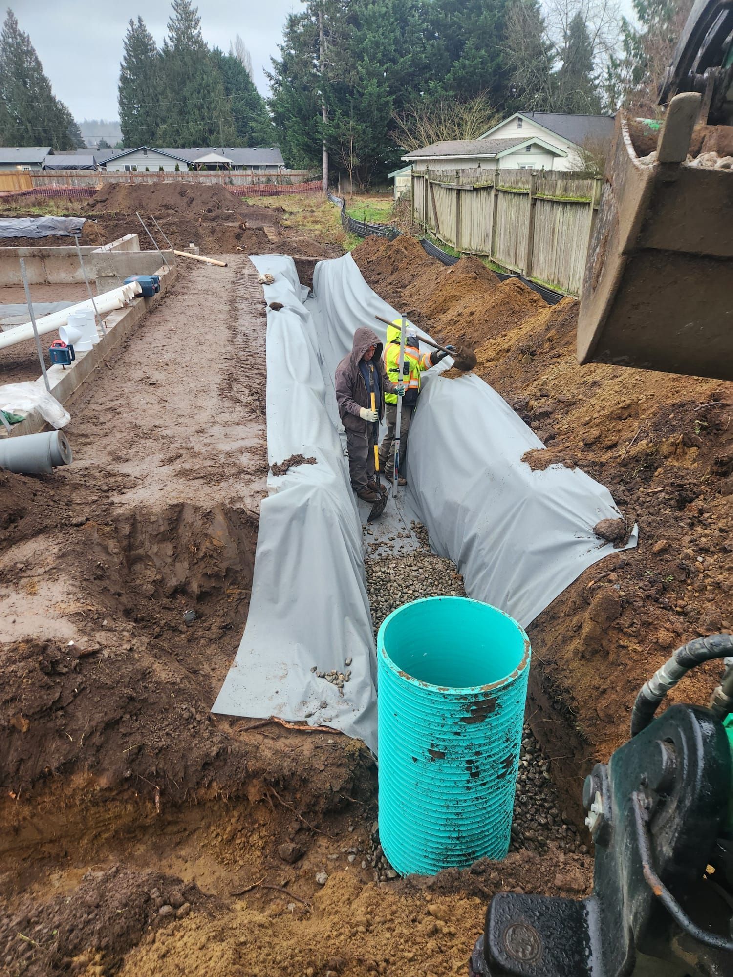 Construction worker in trench with large blue drain pipe, using tools. Grey fabric lines the trench.