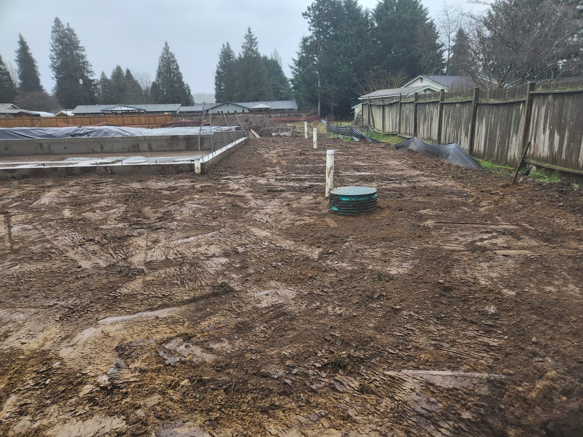 Muddy construction site with partially built foundation, pipes, and wooden fence. Overcast day.