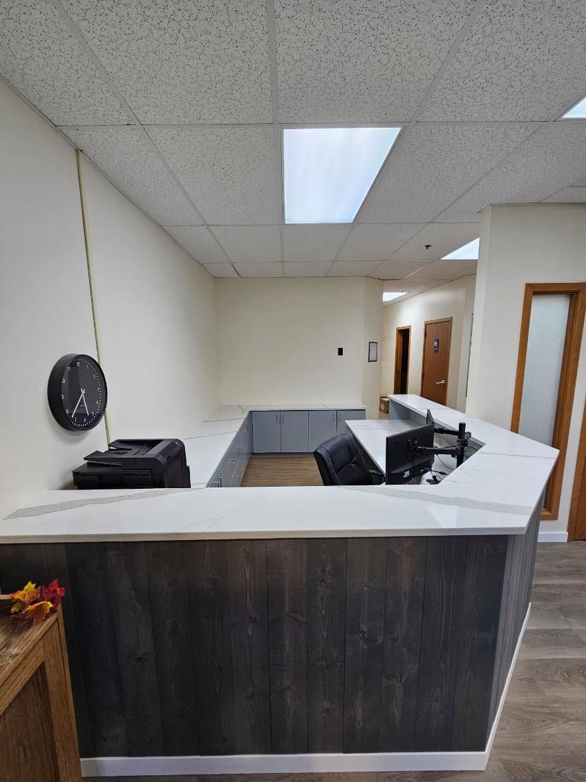Reception desk in an office. White counter with dark wood paneling. Ceiling lights illuminate the hallway.