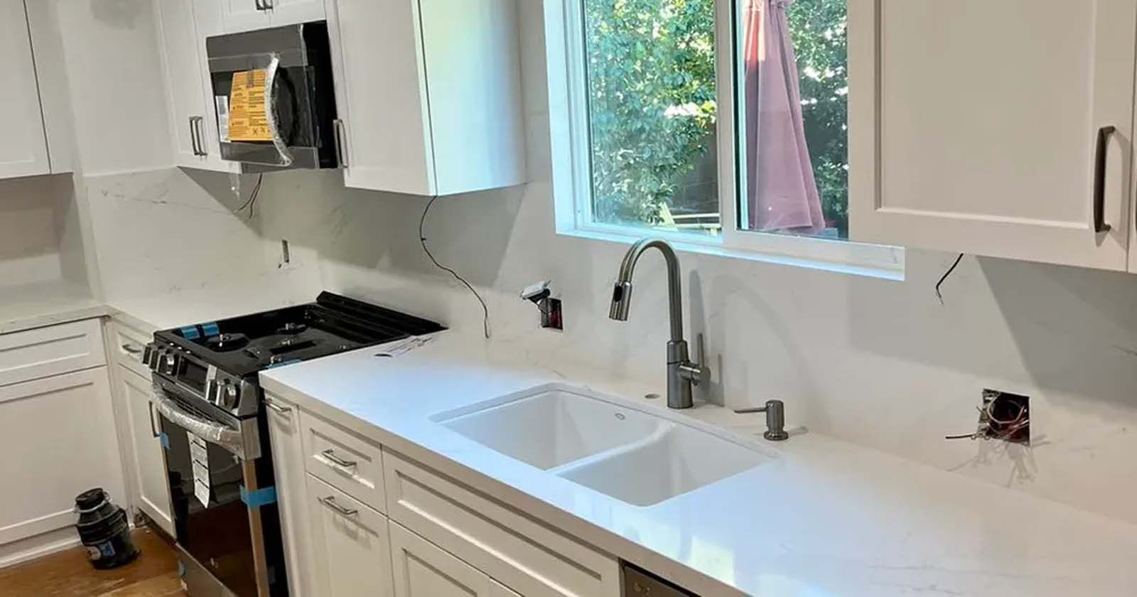 A kitchen with white cabinets , a sink , a stove , and a window.