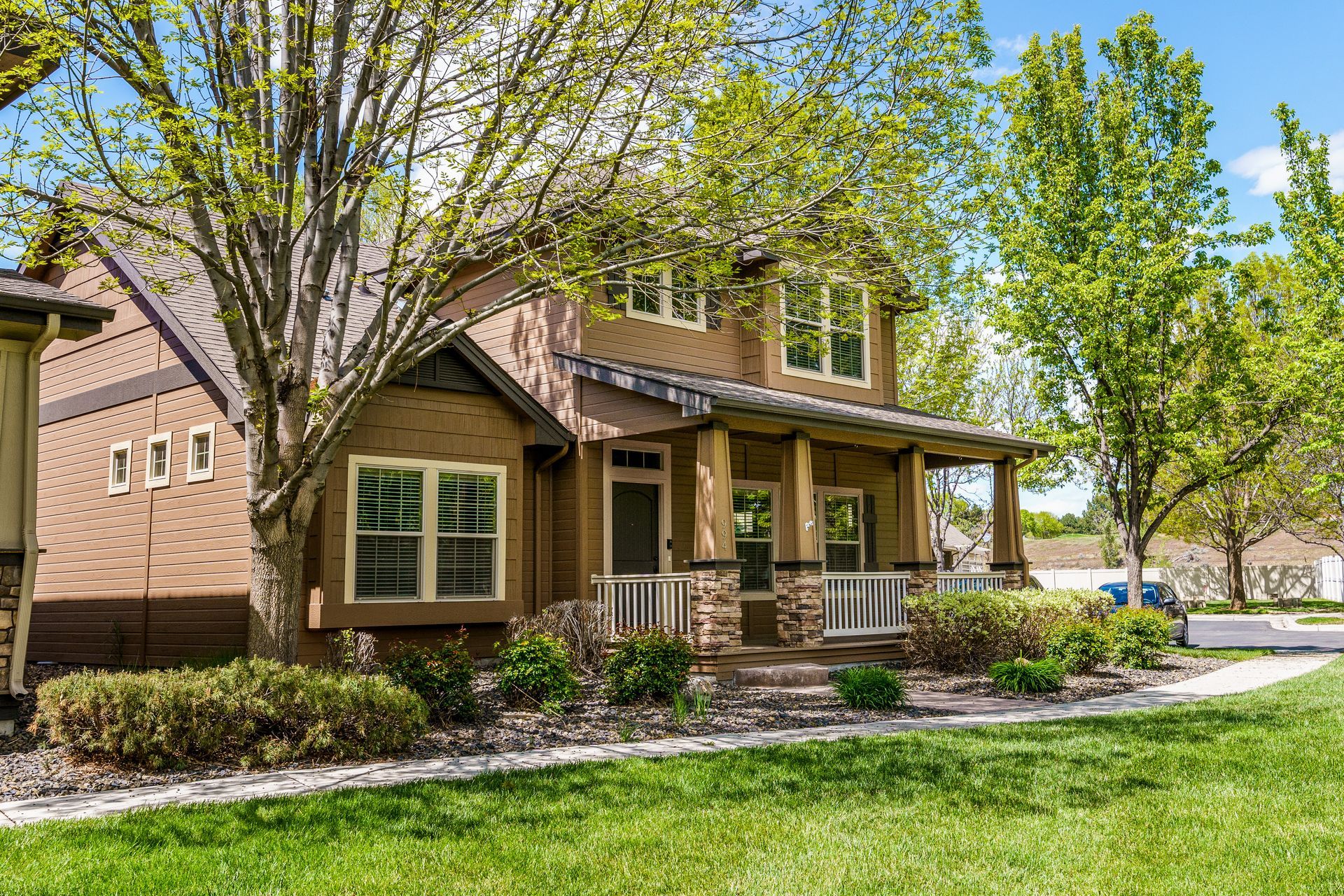 Two-story brown house with a porch and green lawn, trees in the yard under a partly cloudy sky.