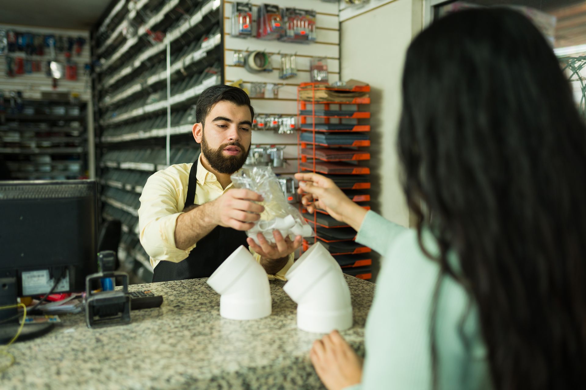 A store clerk hands a customer a bag over the counter, plumbing supplies visible.