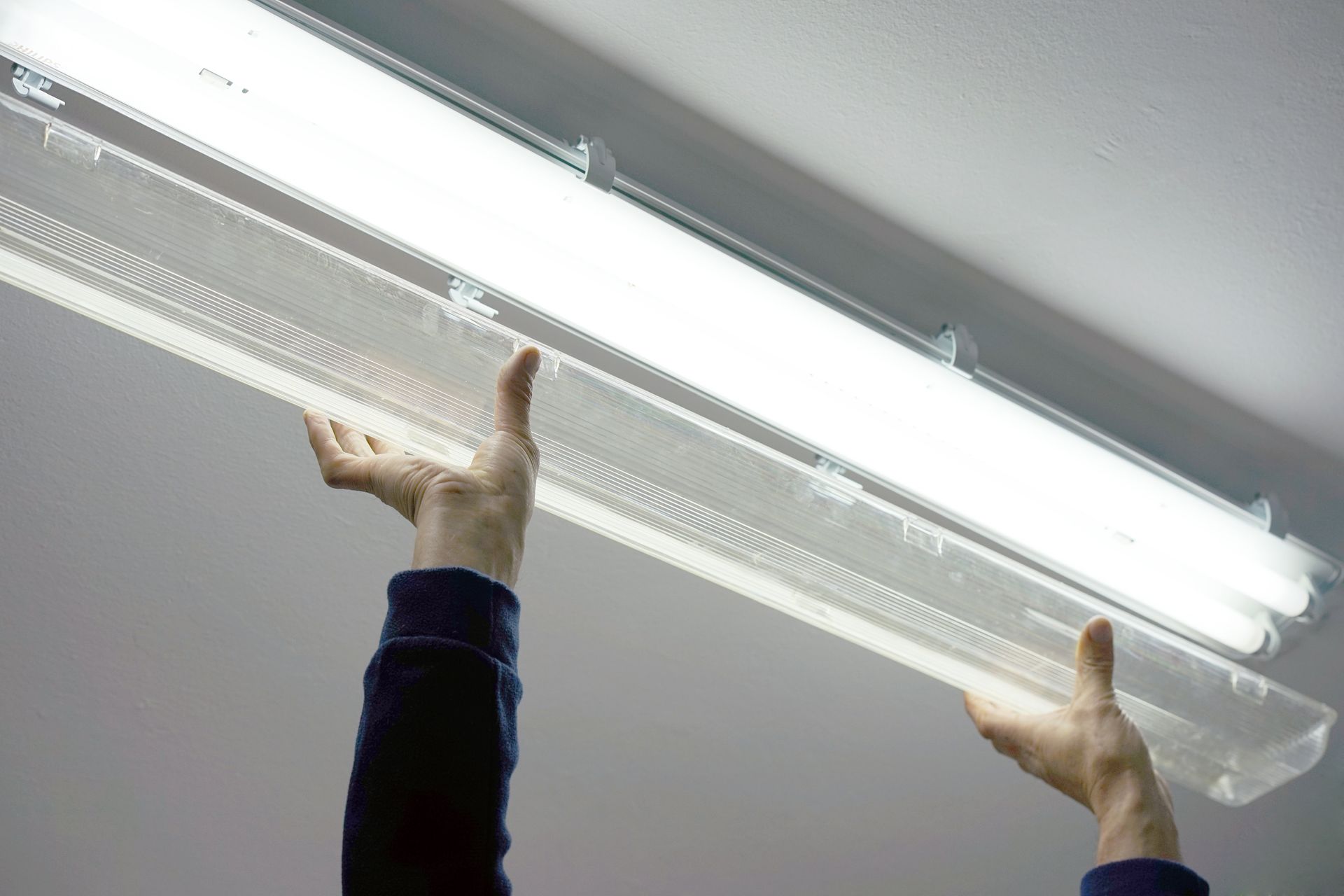 Hands removing a translucent cover from a fluorescent light fixture on a ceiling.