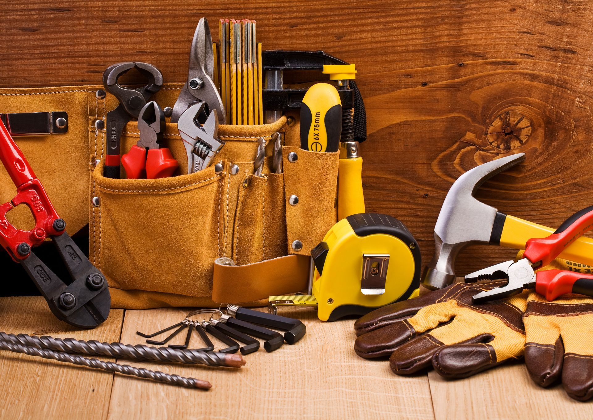 Tool belt with assorted tools, including pliers, hammer, tape measure, and gloves, on a wood surface.