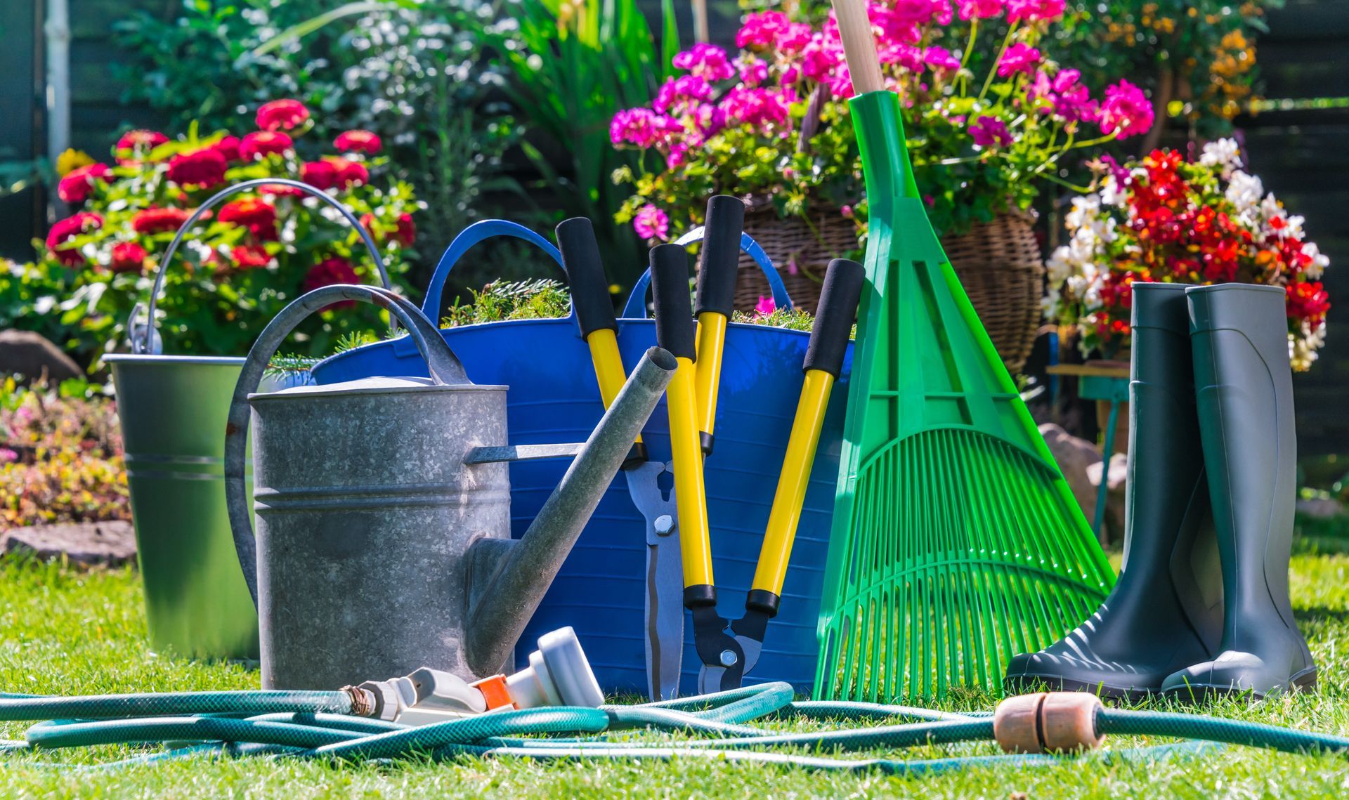 Garden tools, including a watering can, pruners, and rake, rest on a grassy lawn with flowers in the background.