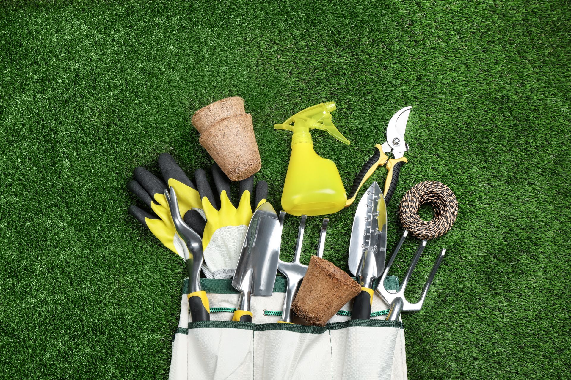Gardening tools in a canvas tool bag on a grassy lawn: gloves, trowels, spray bottle, shears, pots.