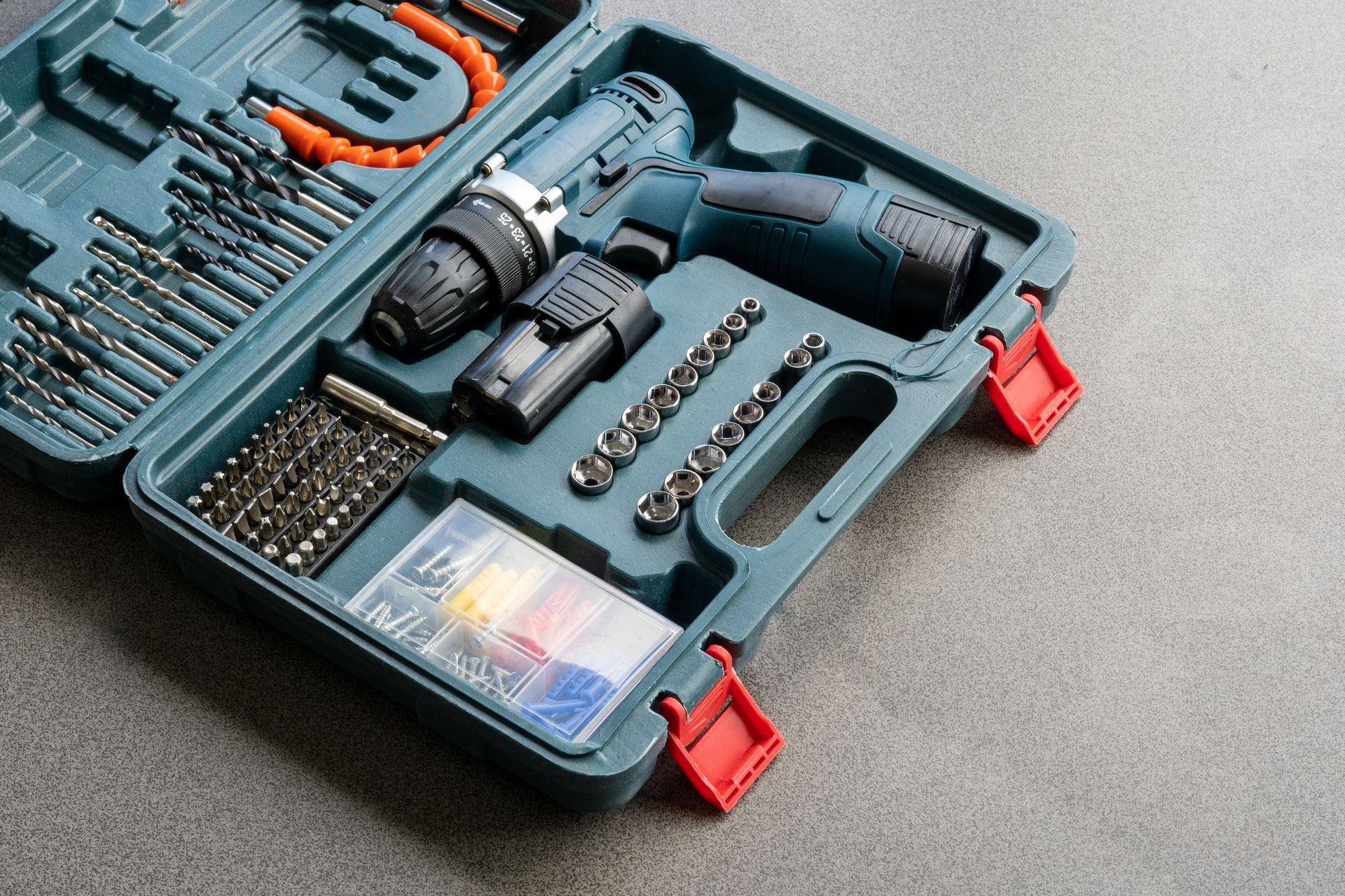 Open toolbox containing a cordless drill, drill bits, and various attachments, in a grey setting.