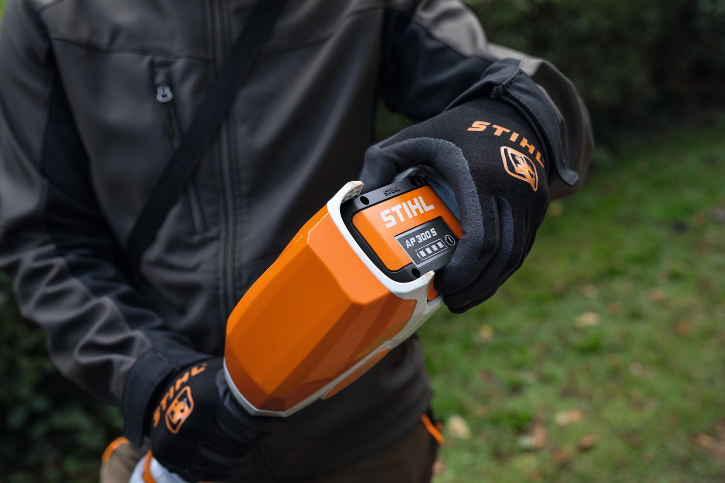 A person in a gray jacket and Stihl gloves inserts a Stihl battery into a handheld outdoor power tool.