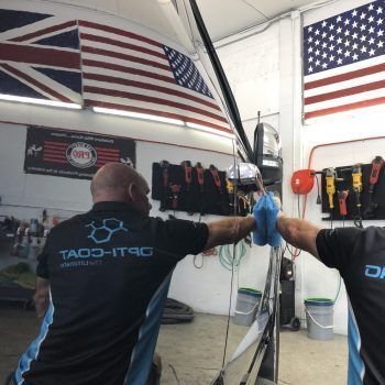 Two men are working on a car in a garage and one of them is wearing a taco-it90 shirt