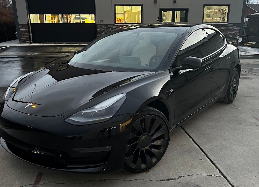 Black Tesla Model 3 parked in front of a building with garage door, windows, and concrete pavement.