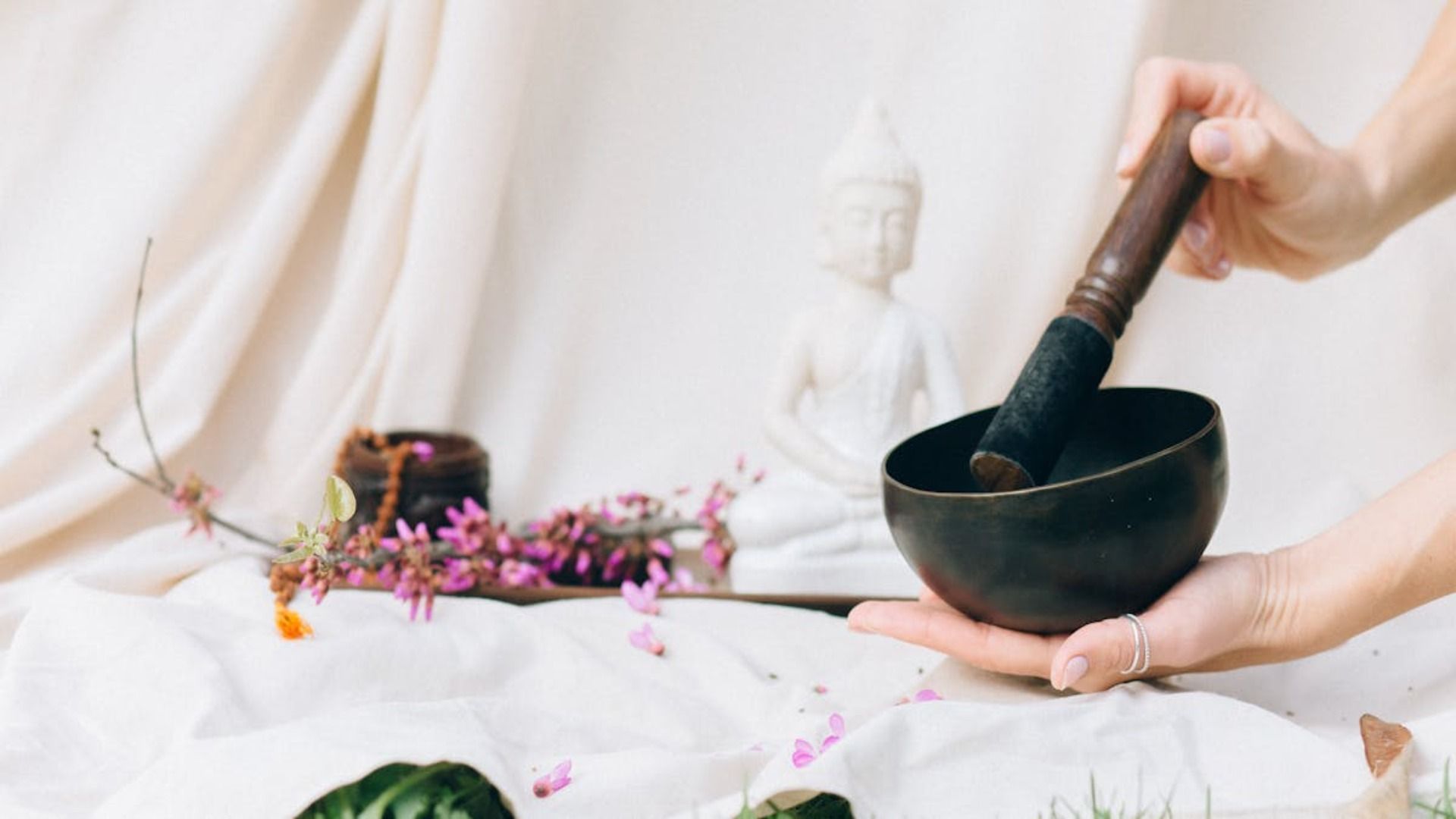 Manos sosteniendo un cuenco cantor con un mazo cerca de una estatua de Buda y flores sobre tela.