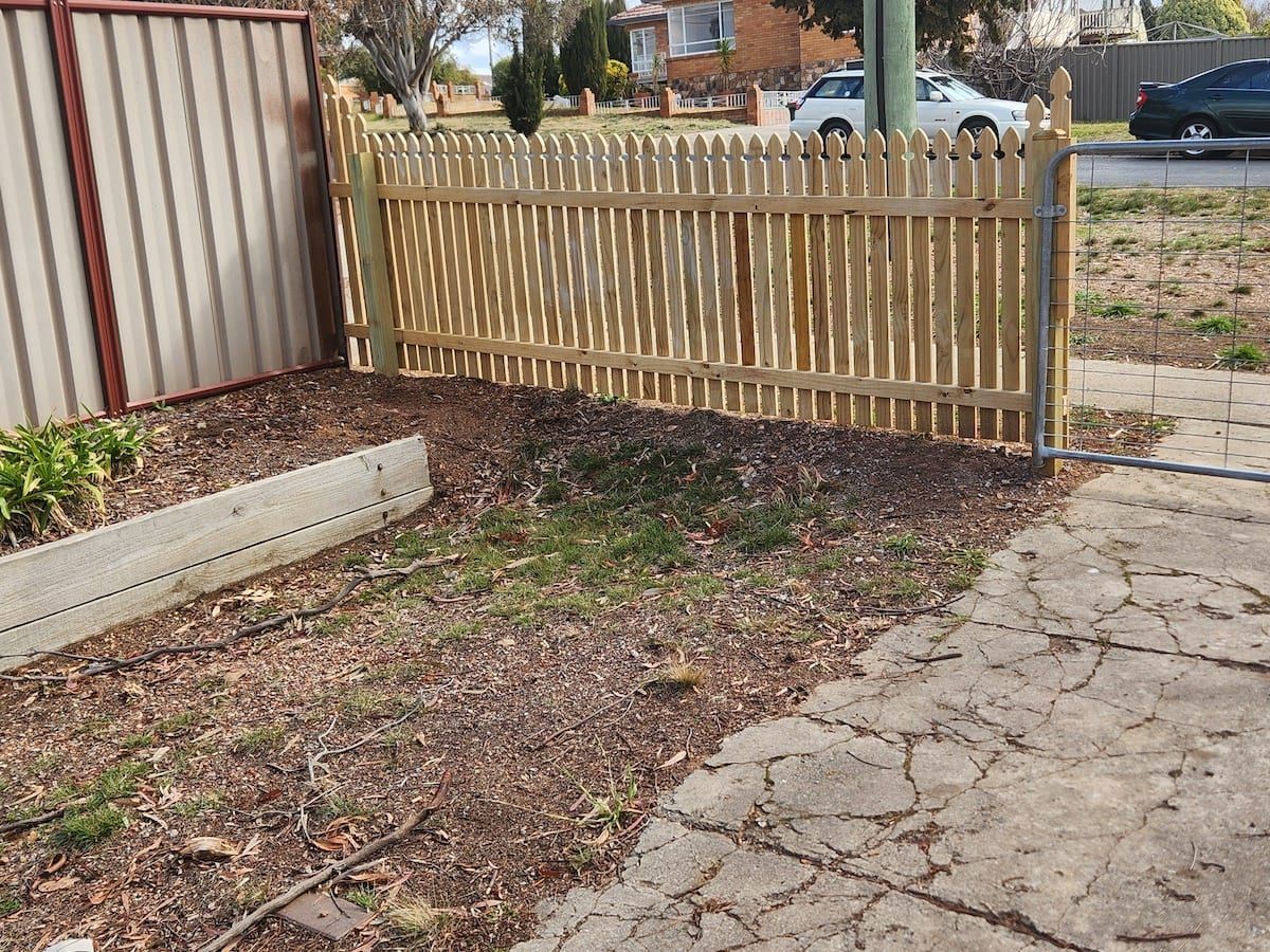 Wooden picket fence in front yard with concrete and dirt.