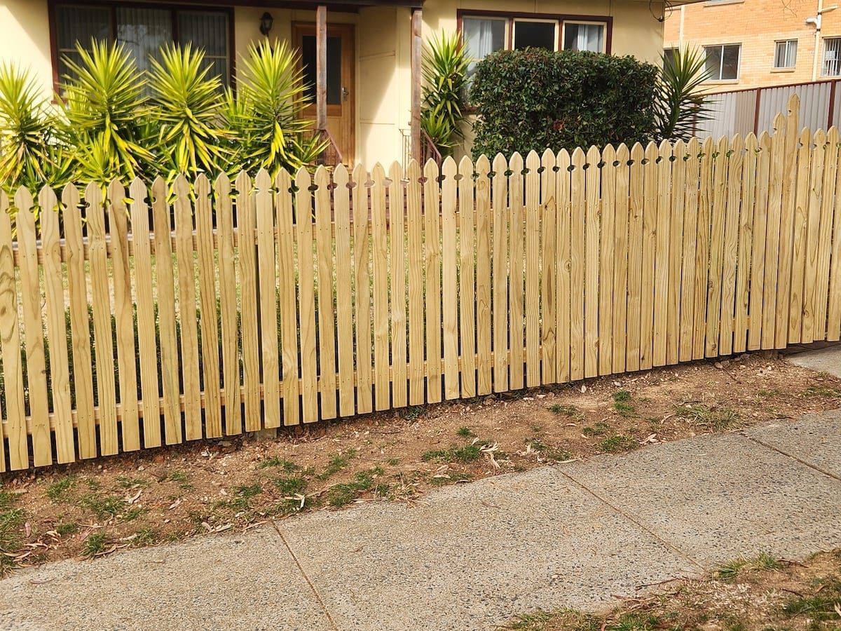 Wooden picket fence in front of a house, beside a sidewalk with dry grass in the foreground.