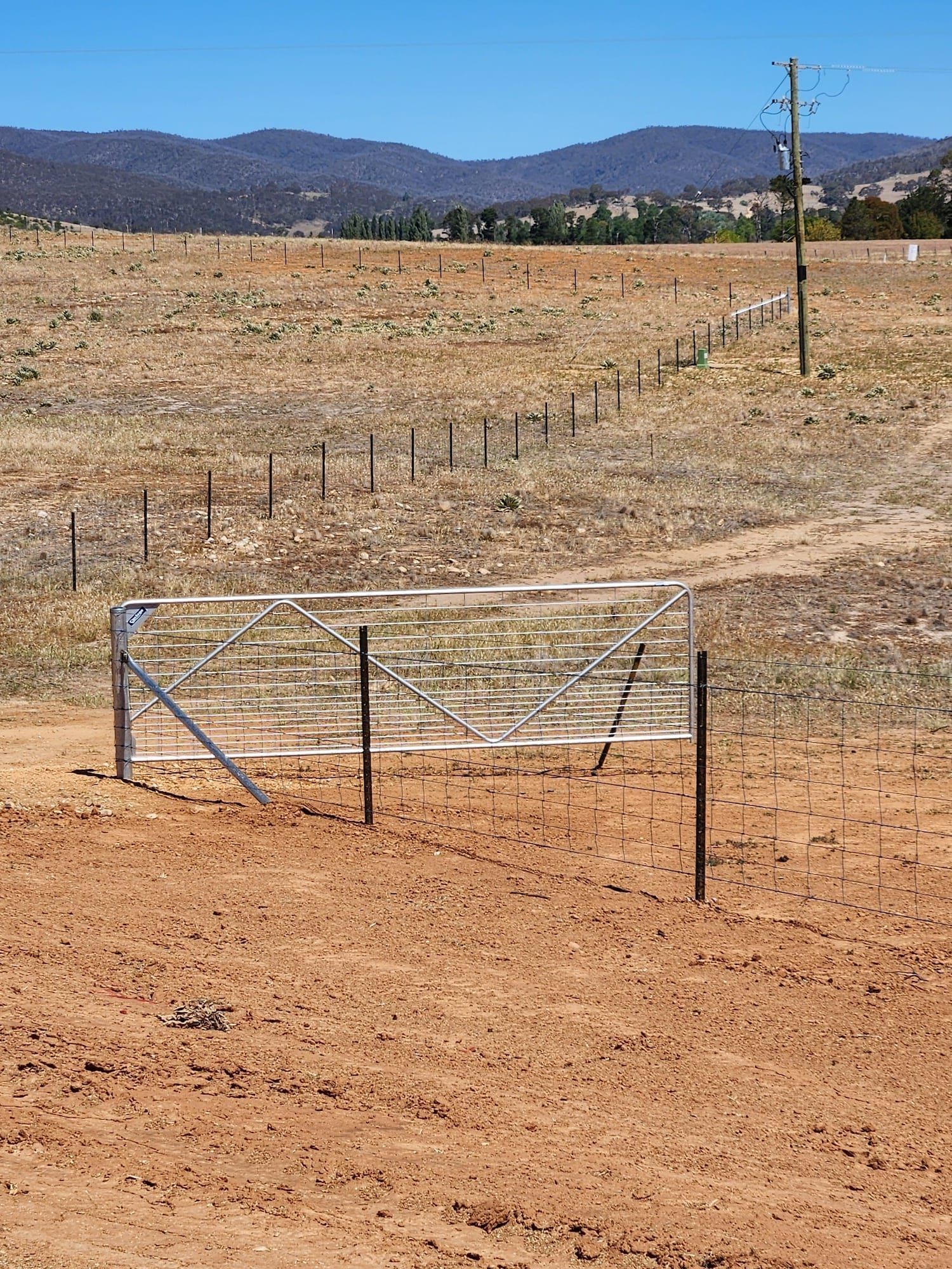 Metal gate in a dry, brown field with a fence leading towards hills under a clear, blue sky.